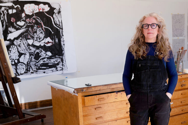 A woman artist stands for a portrait with her hands in her pocket in a studio with a black and white painting in the background and an easel to one side.