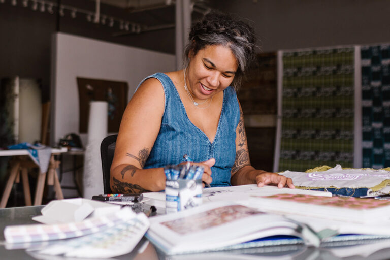 A smiling woman artist works sitting at a table covered in books and supplies.