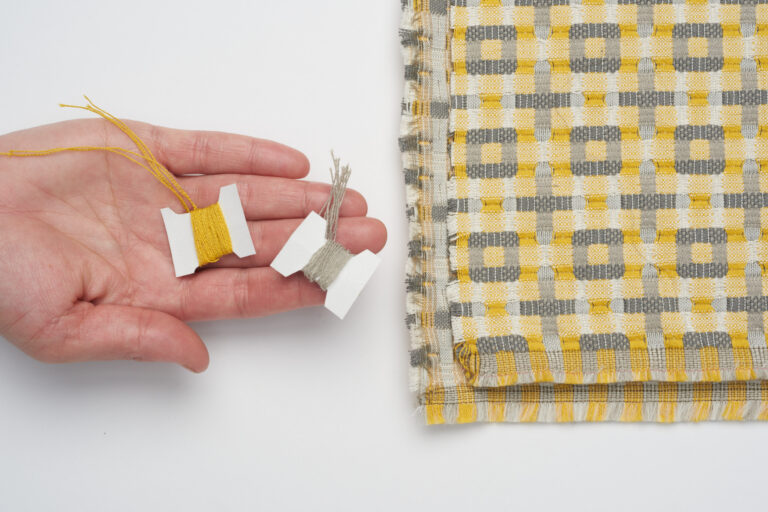 A textiile with a pattern consiting of plus signs and squares on a white background with a hand holding two colored yarn spools.